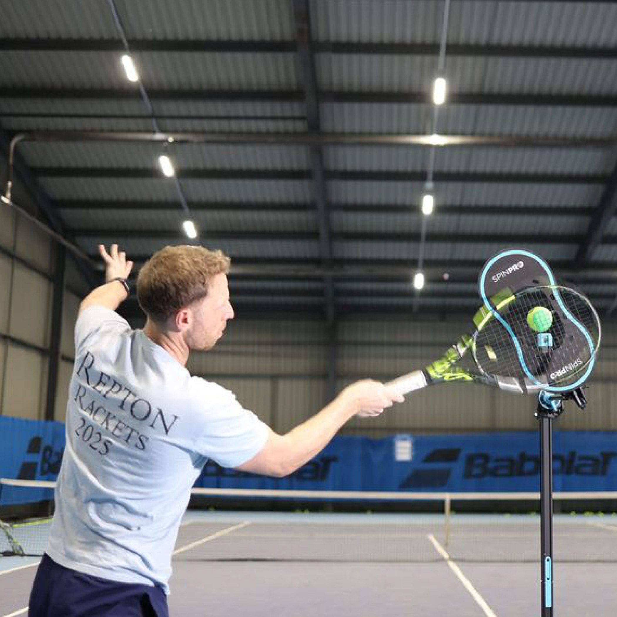 Player driving a high topspin forehand with the SpinPro on an indoor court, viewed from behind
